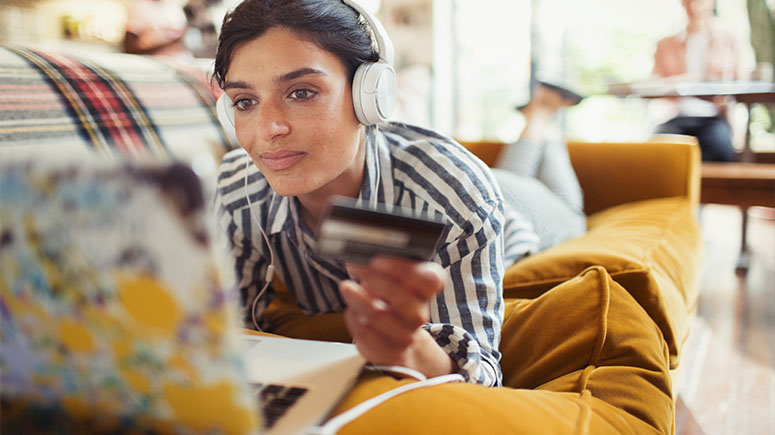 Lady with headphones on laying on a sofa using her card to make a purchase online