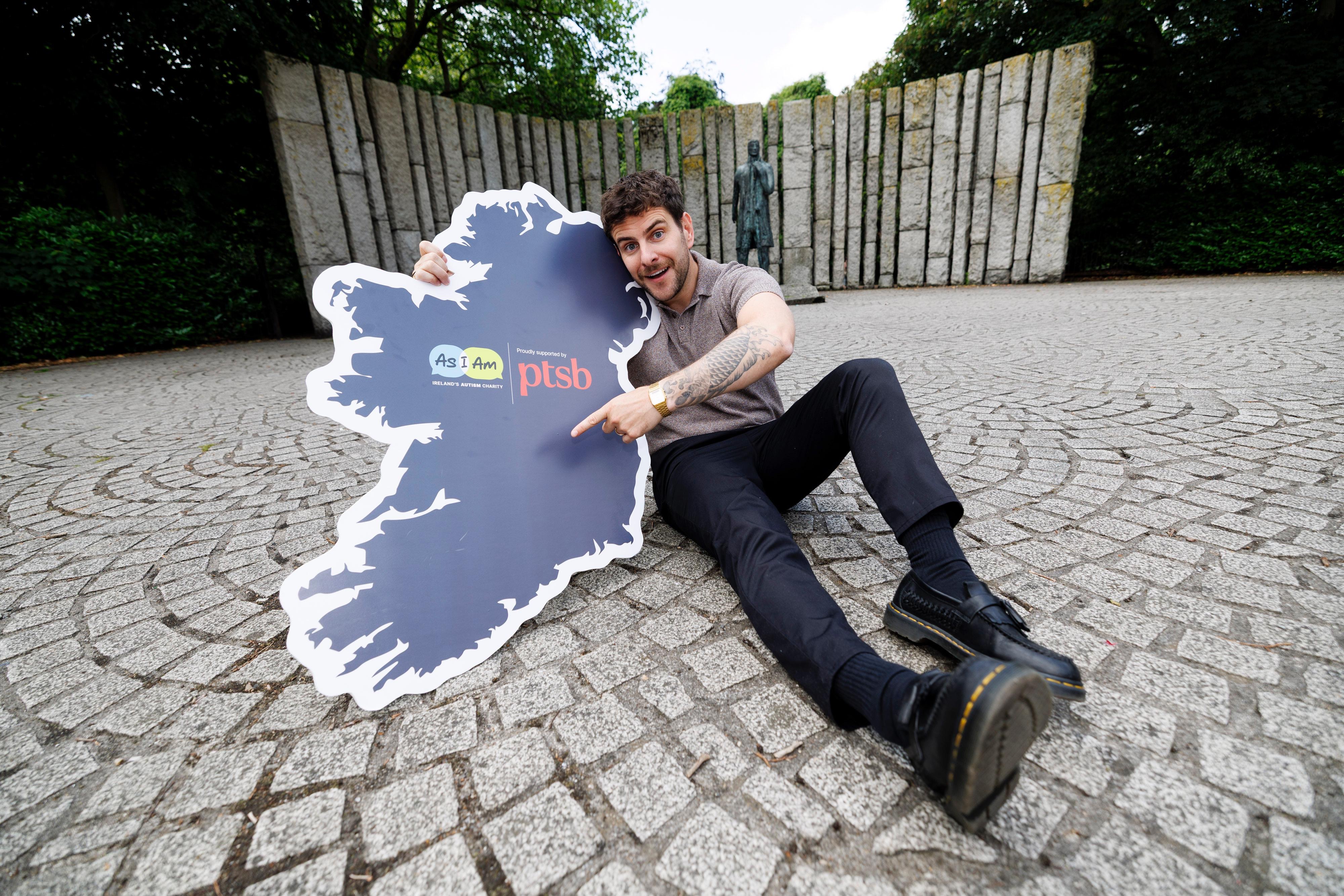 Eric Roberts, sitting in St. Green park, holding a cut out map of Ireland with AsIAm and PTSB logos and pointing to it