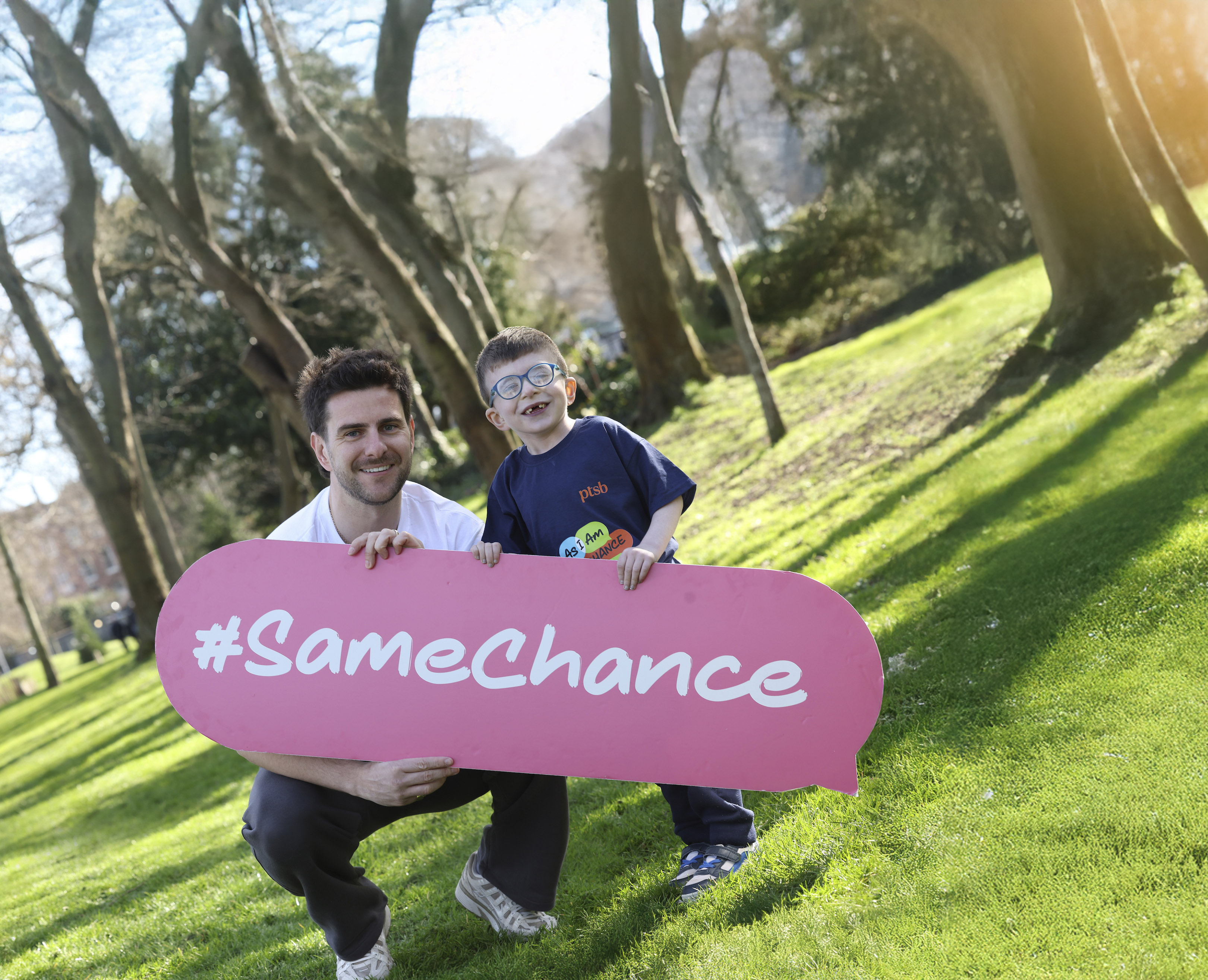A man with a boy holding a banner with text #Same chance, smiling, standing on a hill with trees