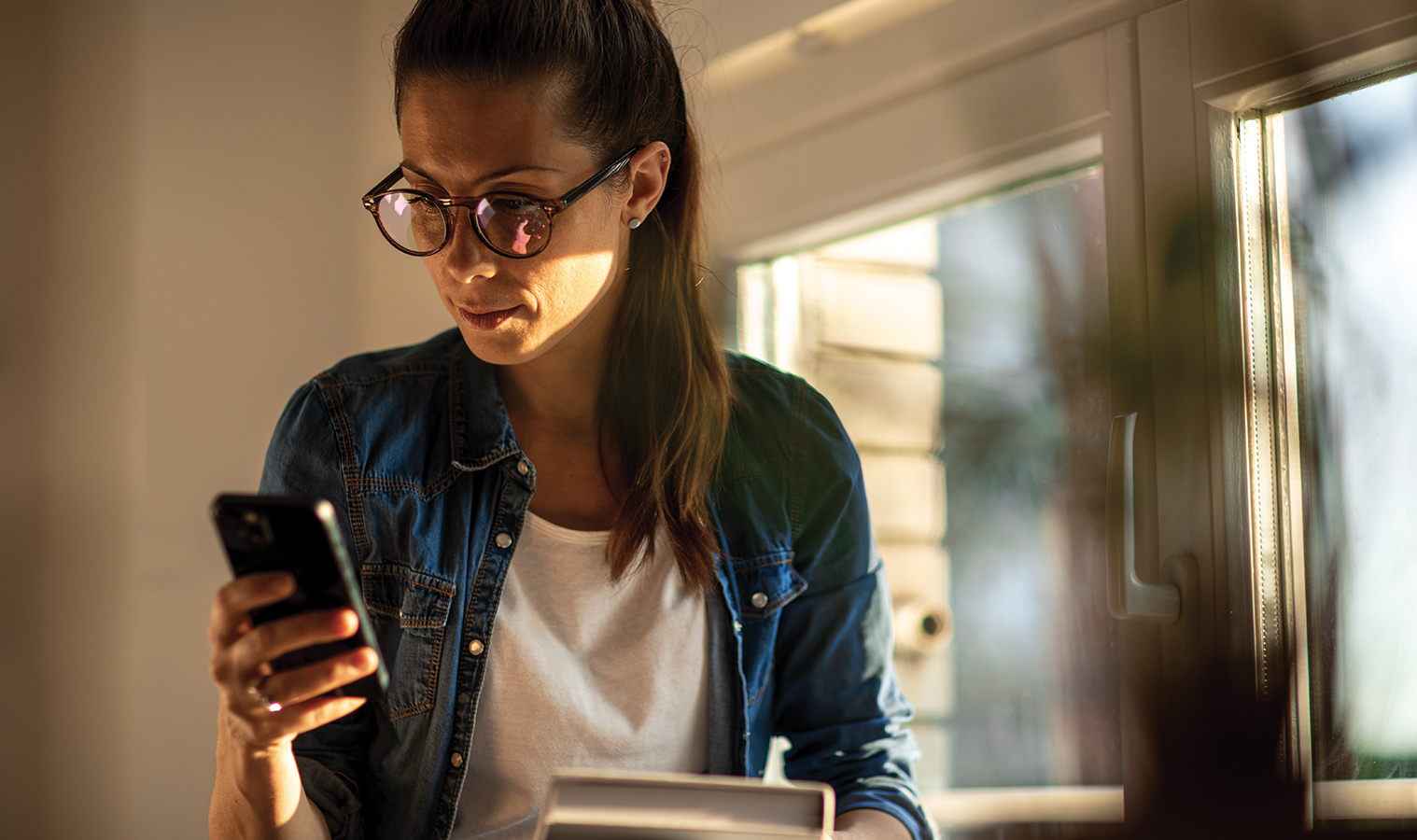 woman in glasses standing next to a window inside a house, looking at her mobile  