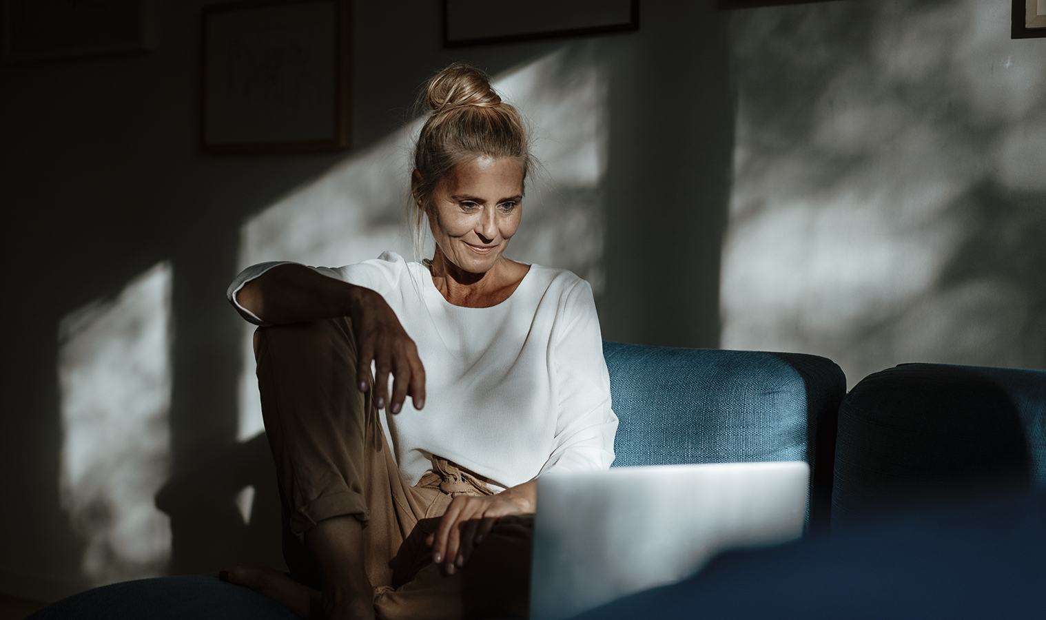 A lady sitting on her sofa looking at a laptop with the sun shining through the window on her face