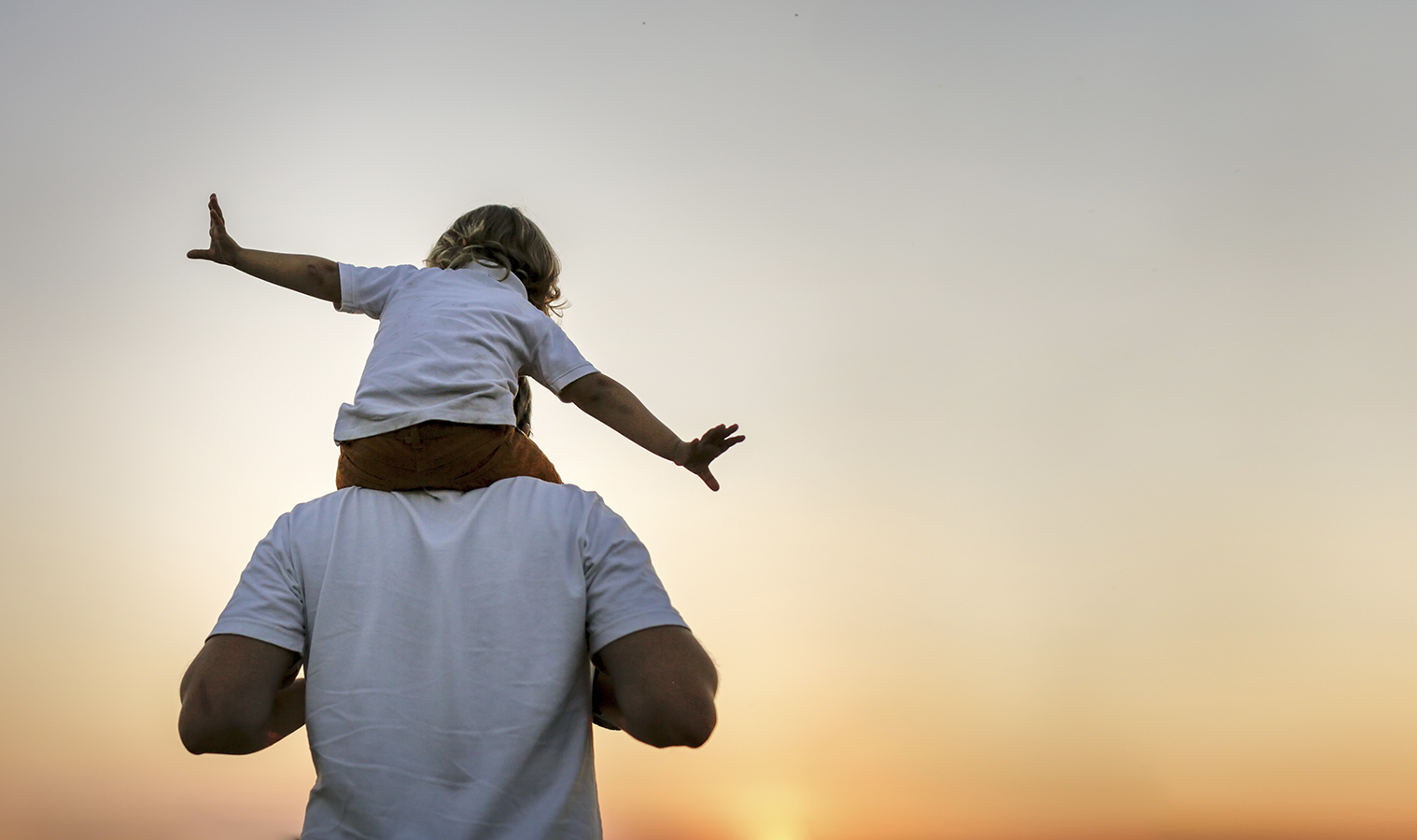 A little boy on his fathers shoulders pretending he is an airplane as they walk towards a sunset