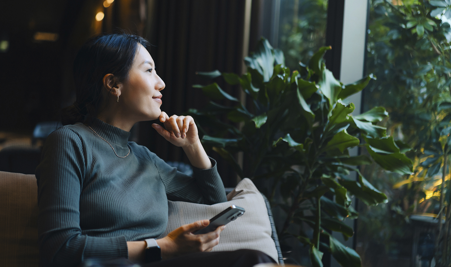 A lady sitting on a sofa with a thinking expression looking out the window with plants surrounding her